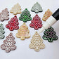 One pair of green, red and gold pearl marble cookie Christmas tree dangle earring blanks paint filled in light pink and white, alongside one unpainted pairs with protective paper attached, with an uncapped Posca paint pen positioned ready to paint fill the engraved details