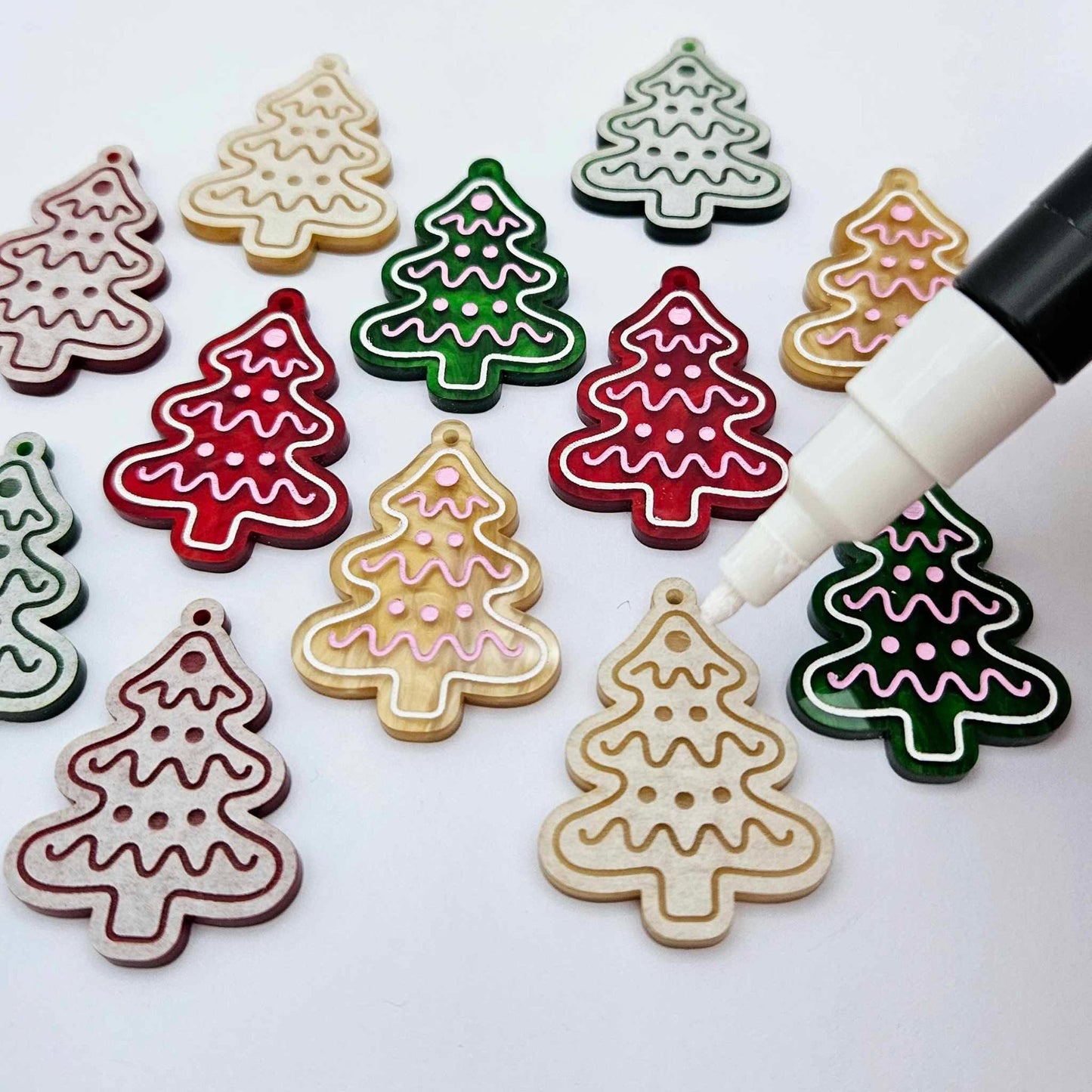 One pair of green, red and gold pearl marble cookie Christmas tree dangle earring blanks paint filled in light pink and white, alongside one unpainted pairs with protective paper attached, with an uncapped Posca paint pen positioned ready to paint fill the engraved details