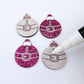 One pair of paint-filled hot pink glitter Santa bauble dangle earring blanks with one unpainted pair with protective paper attached, with an uncapped Posca paint pen positioned ready to paint fill the engraved Santa belt details
