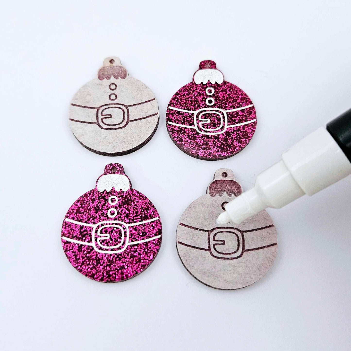 One pair of paint-filled hot pink glitter Santa bauble dangle earring blanks with one unpainted pair with protective paper attached, with an uncapped Posca paint pen positioned ready to paint fill the engraved Santa belt details