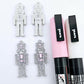 A pair of 40mm nutcracker Christmas dangle earring blanks in silver glitter acrylic with engraved detail paint filled white and pink and a pair not painted