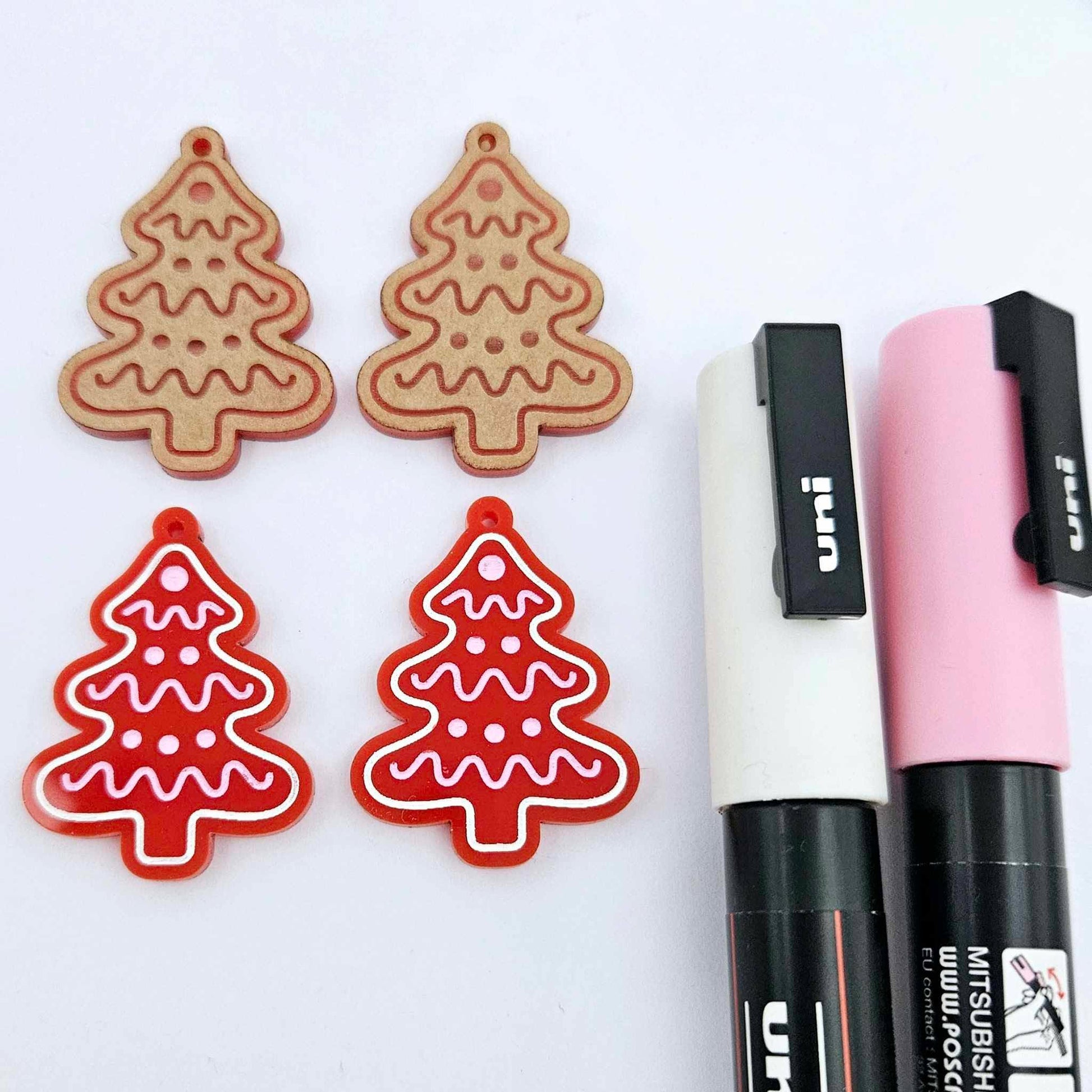 One pair of solid red cookie Christmas tree dangle earring blanks paint filled in light pink and white, alongside one unpainted pair with protective paper still attached, pictured with Posca paint pens