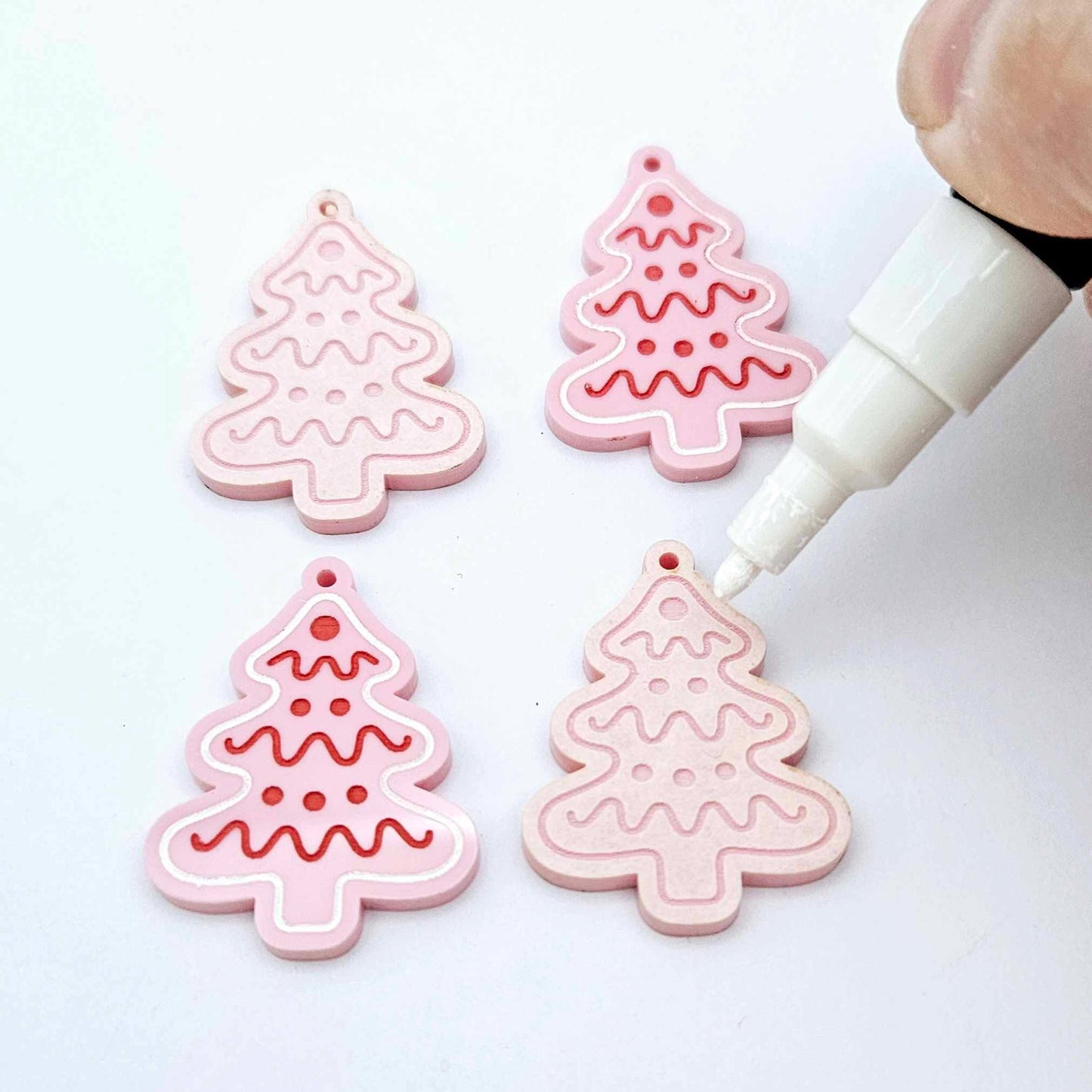 One pair of paint-filled pastel pink cookie Christmas tree dangle earring blanks with one unpainted pair with protective paper attached, with an uncapped Posca paint pen positioned ready to paint fill the engraved details