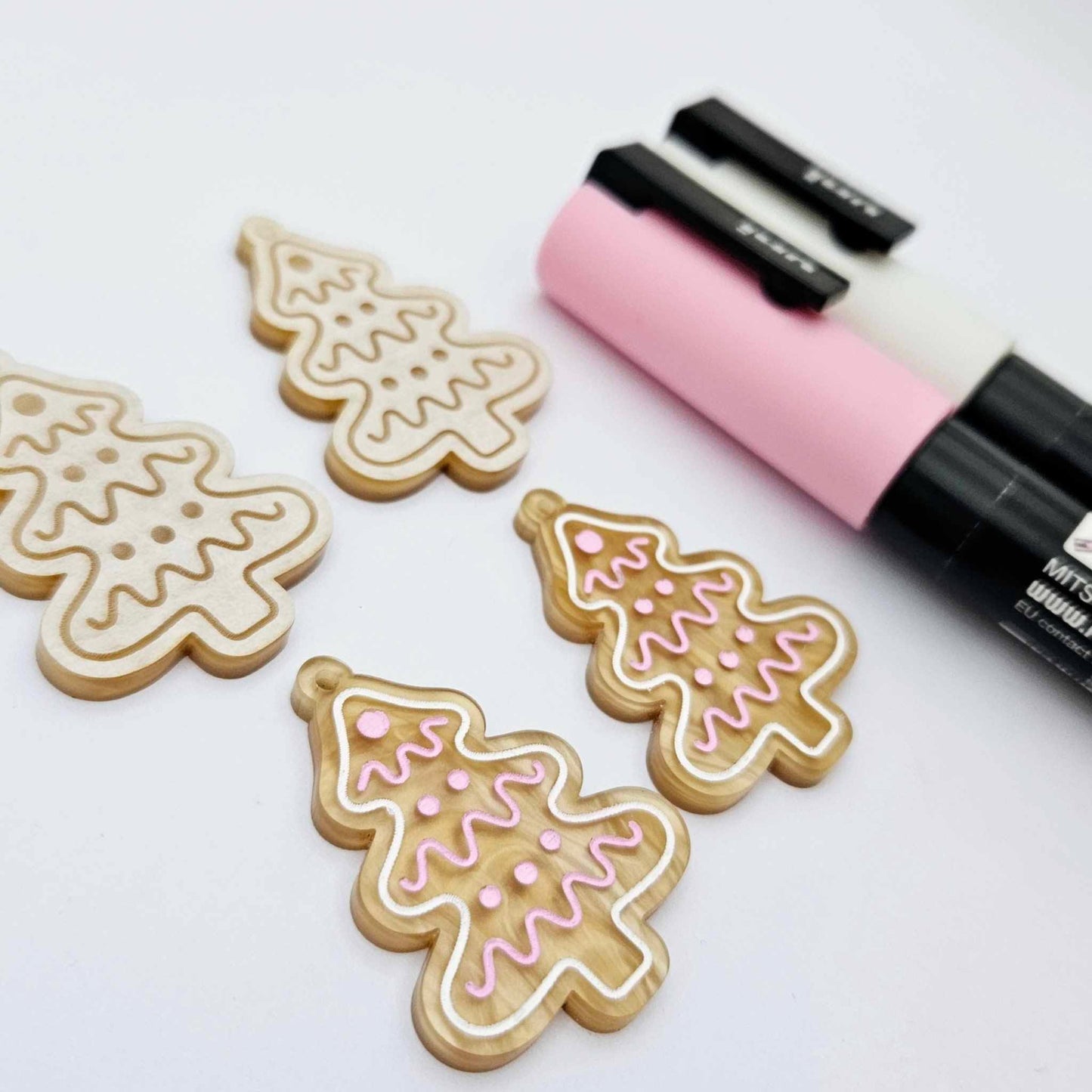 One pair of gold pearl marble cookie Christmas tree dangle earring blanks paint filled in light pink and white, alongside one unpainted pair with protective paper attached, pictured with Posca paint pens