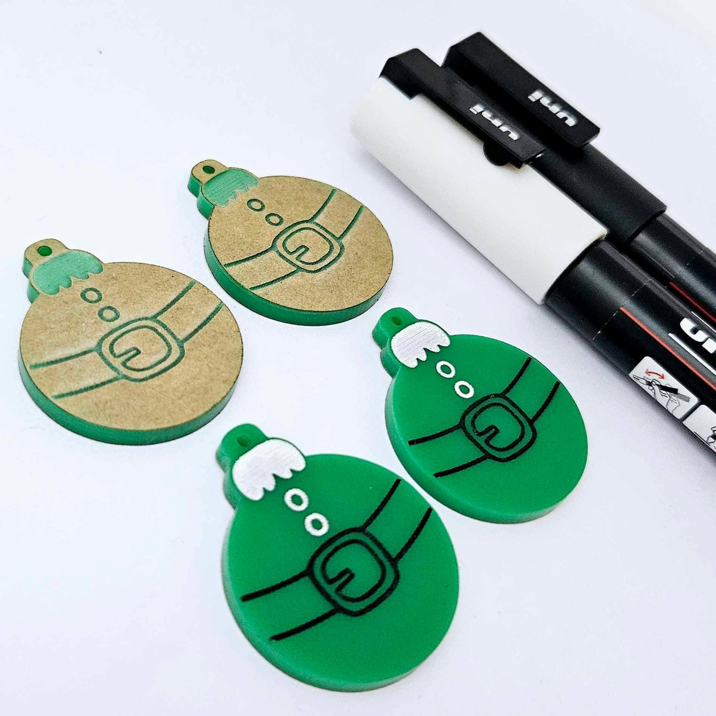 One pair of solid green Santa bauble dangle blanks paint filled in black and white, alongside one unpainted pair with protective paper attached, pictured with Posca paint pens