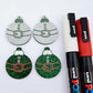 One pair of green glitter Santa bauble dangle blanks paint filled red and white, alongside one unpainted pair with protective paper attached, pictured with Posca paint pens