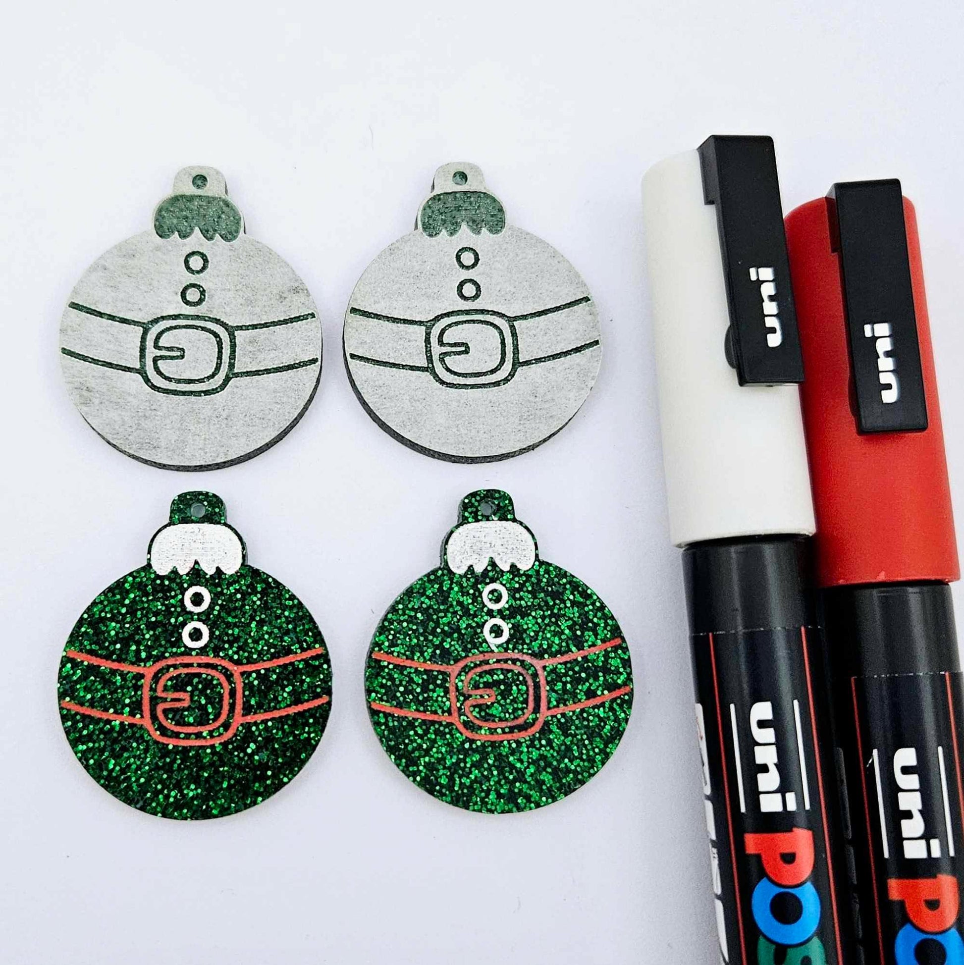 One pair of green glitter Santa bauble dangle blanks paint filled red and white, alongside one unpainted pair with protective paper attached, pictured with Posca paint pens