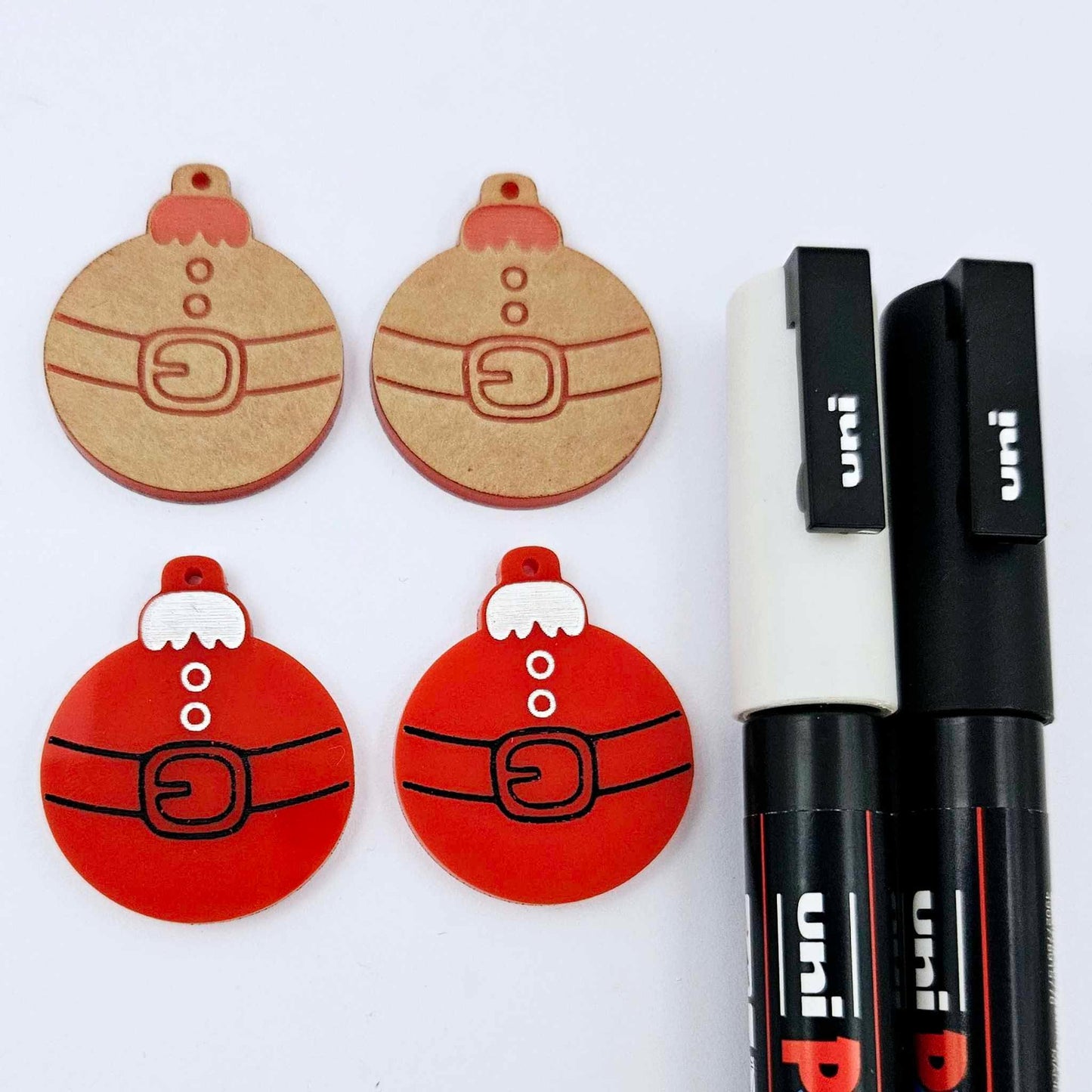 One pair of solid red Santa bauble dangle earring blanks paint filled in black and white, alongside one unpainted pair with protective paper attached, pictured with Posca paint pens