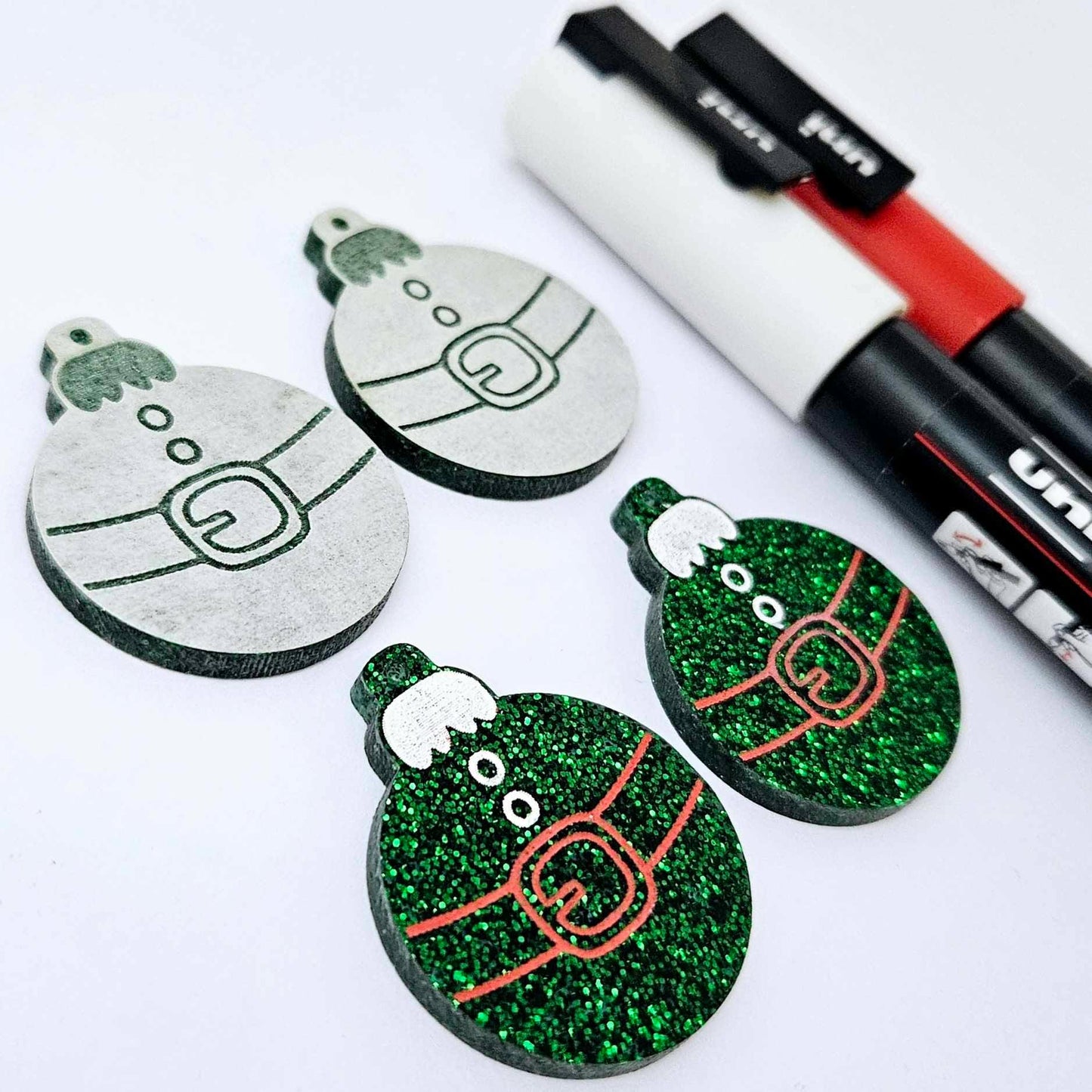 One pair of green glitter Santa bauble dangle blanks paint filled red and white, alongside one unpainted pair with protective paper attached, pictured with Posca paint pens