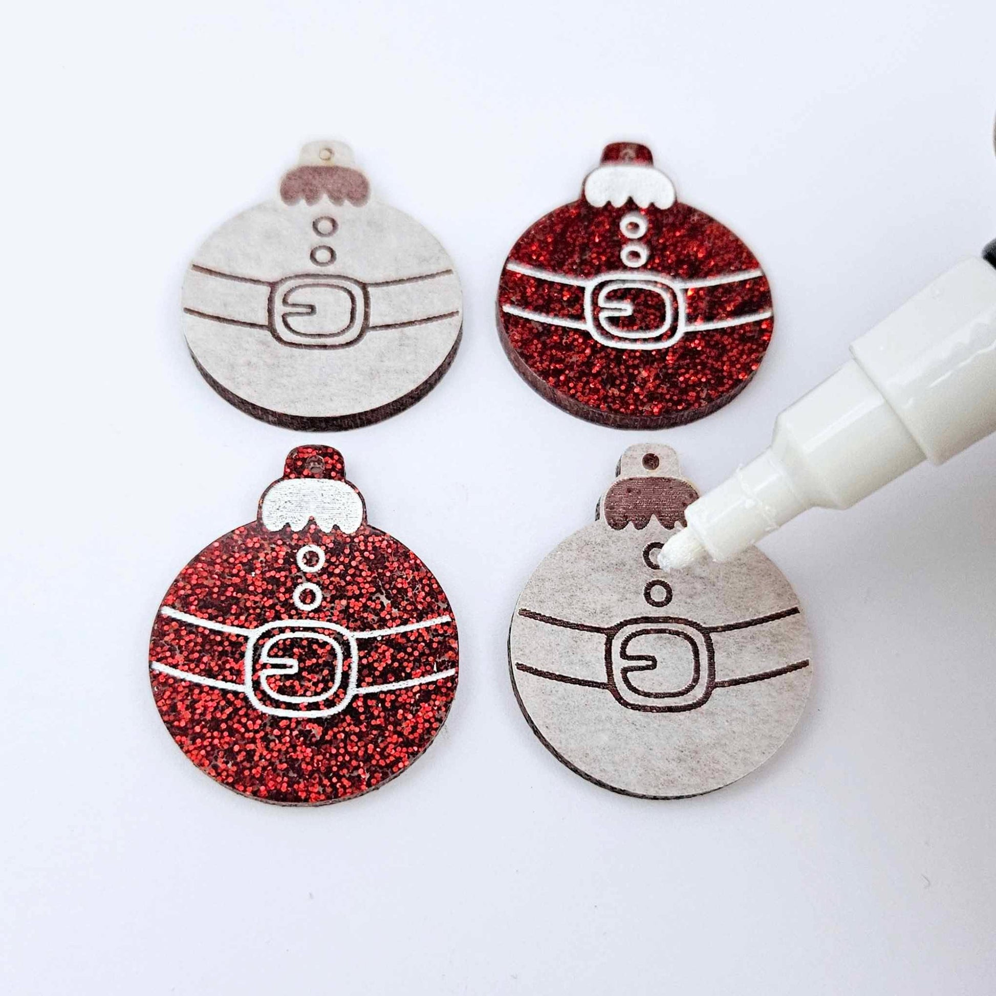 One pair of paint-filled red glitter Santa bauble dangle blanks beside one unpainted pair with protective paper attached, with an uncapped Posca paint pen positioned ready to paint fill the engraved Santa belt details