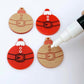 One pair of paint-filled solid red Santa bauble dangle blanks beside one unpainted pair with protective paper attached, with an uncapped Posca paint pen positioned ready to paint fill the engraved Santa belt details
