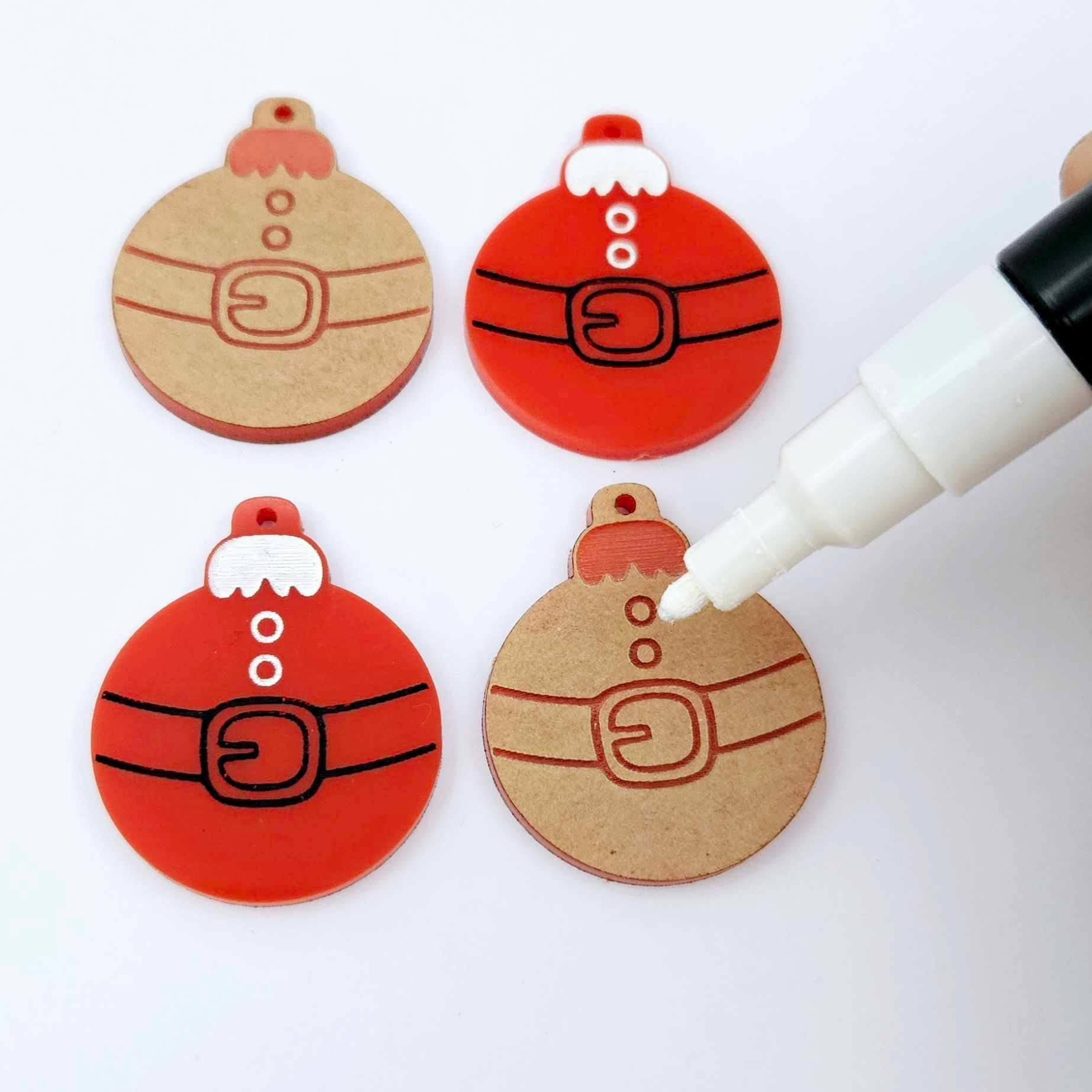 One pair of paint-filled solid red Santa bauble dangle blanks beside one unpainted pair with protective paper attached, with an uncapped Posca paint pen positioned ready to paint fill the engraved Santa belt details