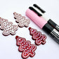 One pair of deep red marble cookie Christmas tree dangle blanks paint filled in light pink and white, alongside one unpainted pair with protective paper attached, pictured with Posca paint pens