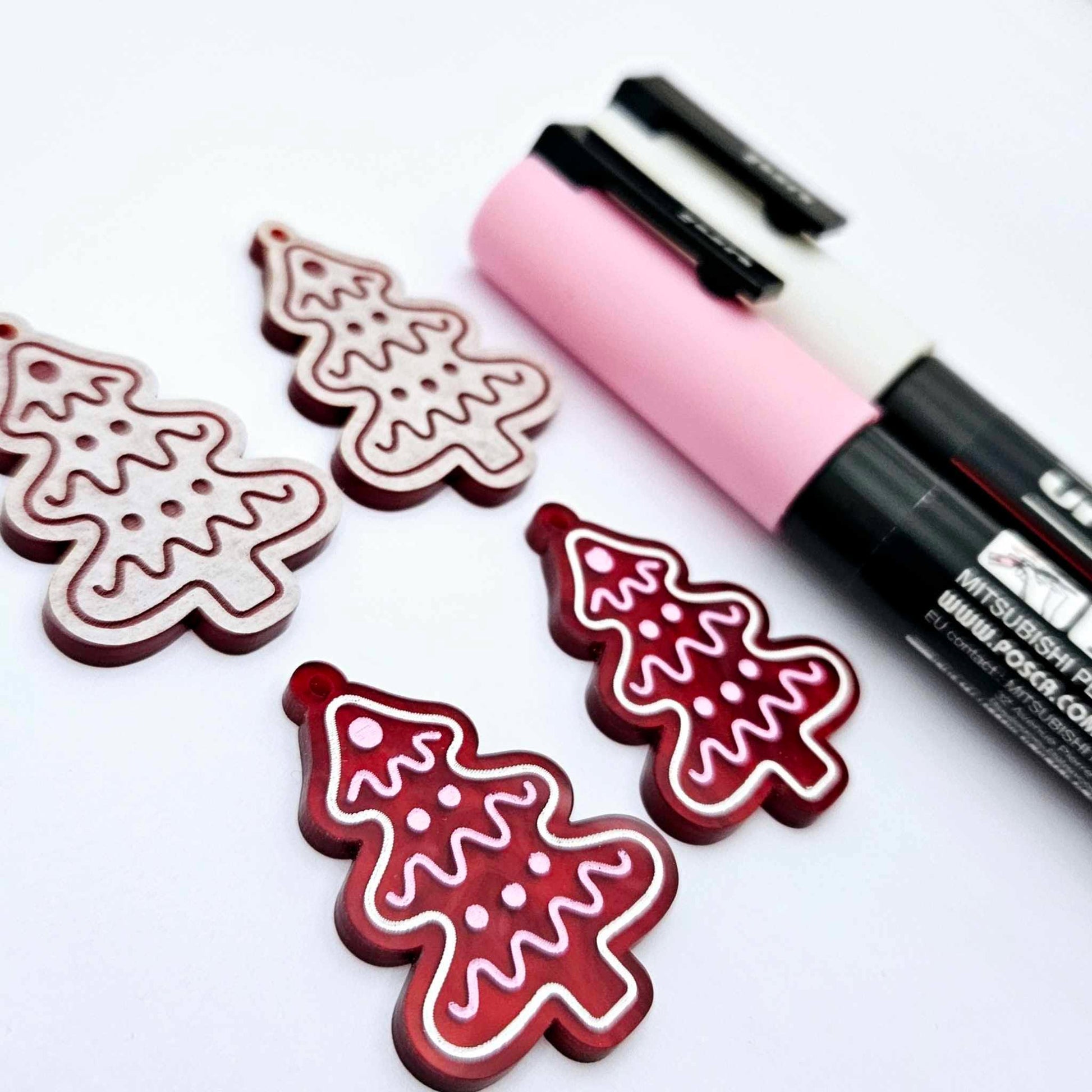One pair of deep red marble cookie Christmas tree dangle blanks paint filled in light pink and white, alongside one unpainted pair with protective paper attached, pictured with Posca paint pens