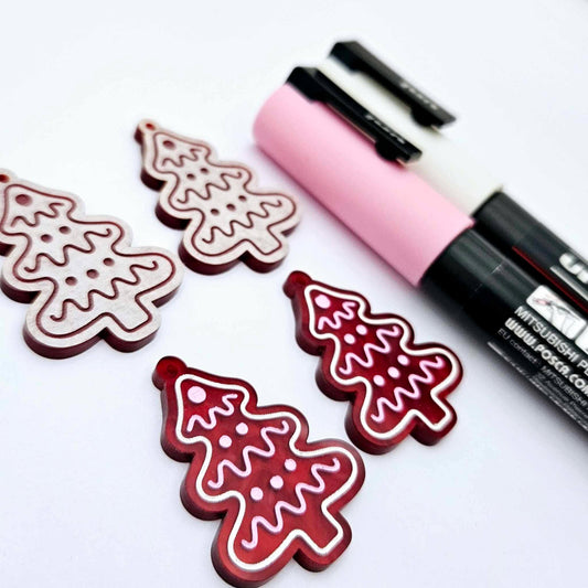 One pair of deep red marble cookie Christmas tree dangle blanks paint filled in light pink and white, alongside one unpainted pair with protective paper attached, pictured with Posca paint pens