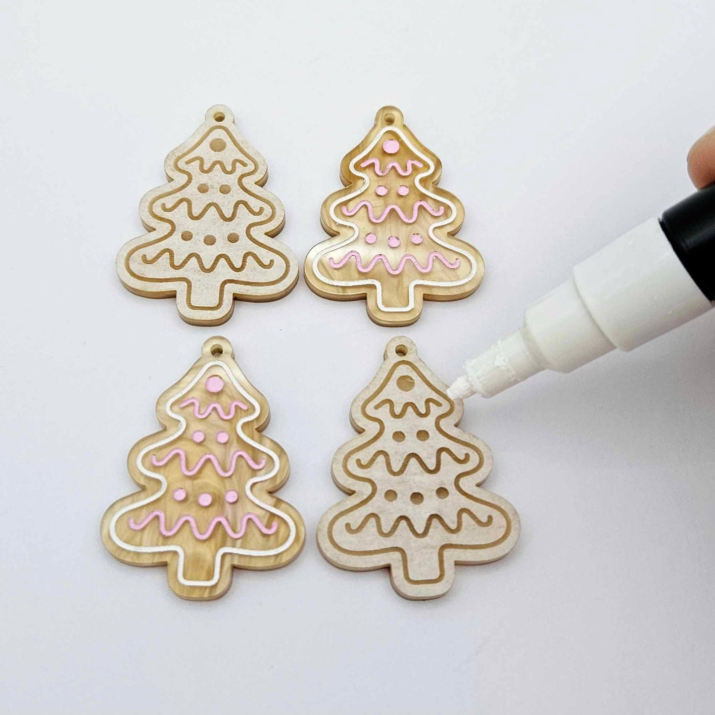 One pair of paint-filled gold pearl marble cookie Christmas tree dangle earring blanks beside one unpainted pair with protective paper attached, with an uncapped Posca paint pen positioned ready to paint fill the engraved details