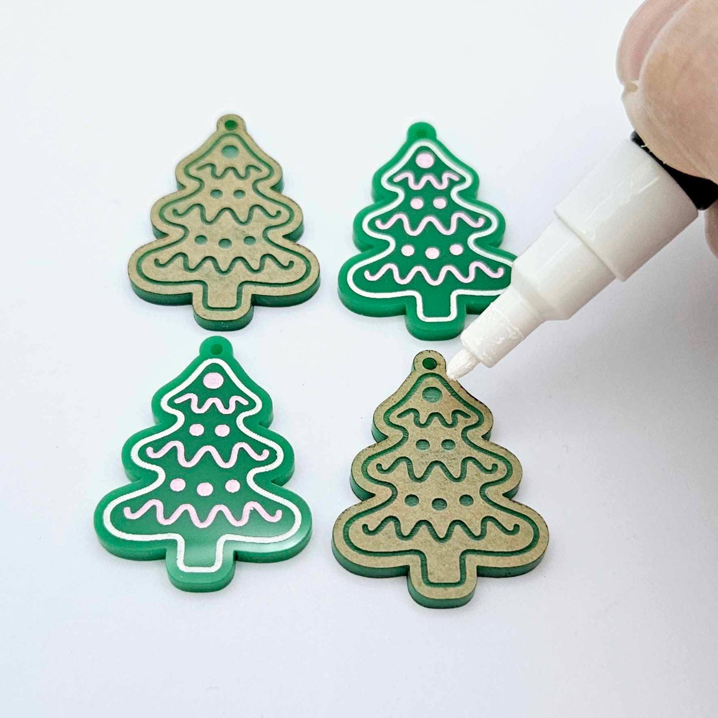 One pair of paint-filled solid green cookie Christmas tree dangle earring blanks with one unpainted pair with protective paper attached, with an uncapped Posca paint pen positioned ready to paint fill the engraved details