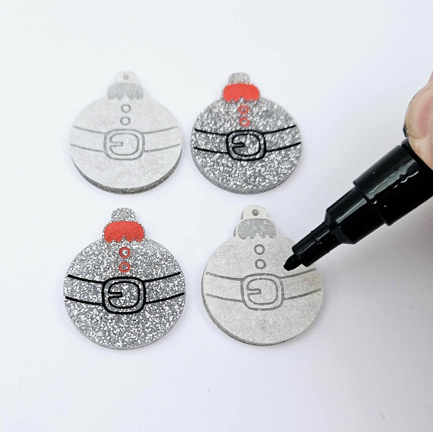 One pair of paint-filled silver glitter Santa bauble dangle blanks with one unpainted pair with protective paper attached, with an uncapped Posca paint pen positioned ready to paint fill the engraved Santa belt details