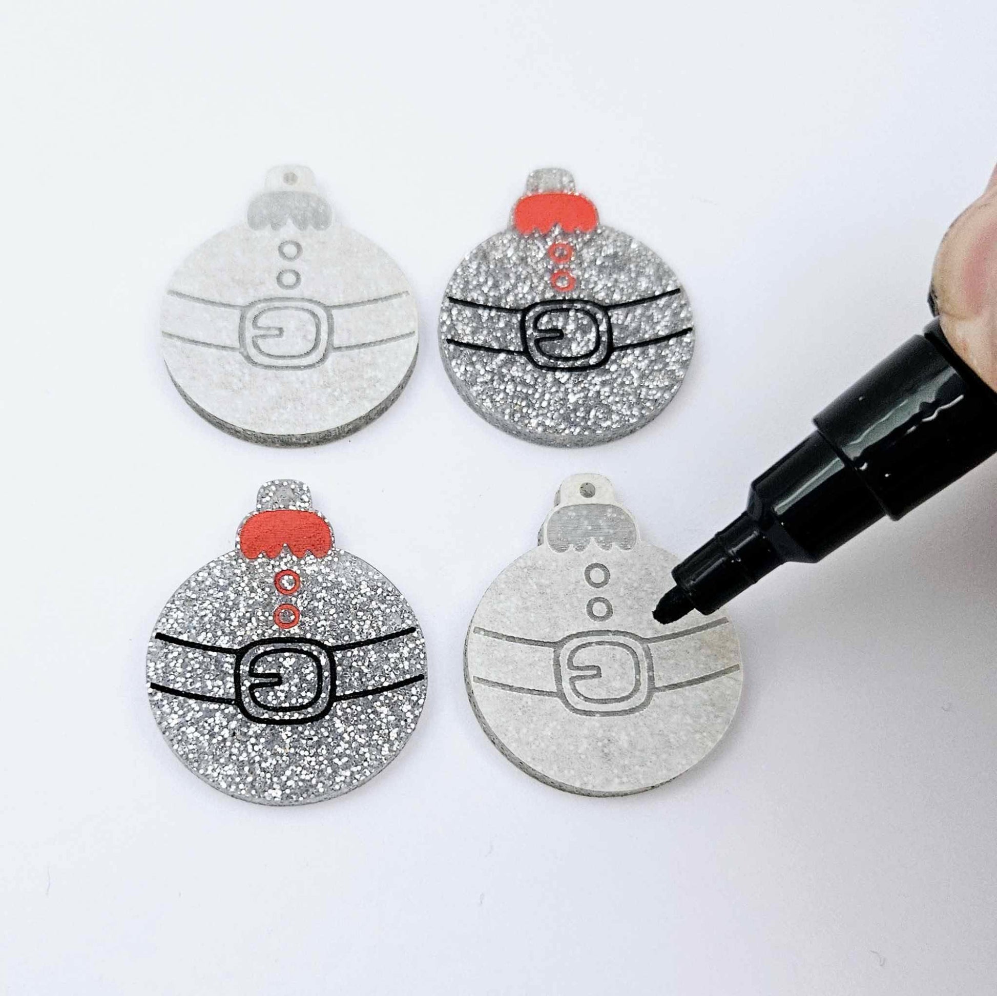 One pair of paint-filled silver glitter Santa bauble dangle blanks with one unpainted pair with protective paper attached, with an uncapped Posca paint pen positioned ready to paint fill the engraved Santa belt details