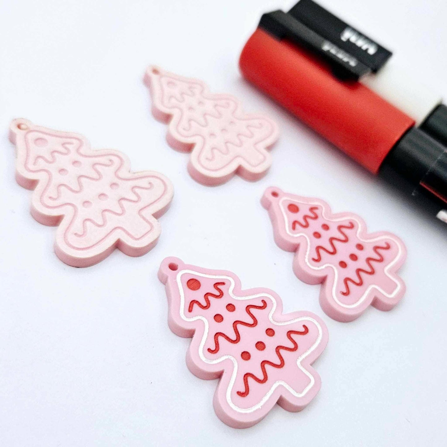 One pair of pastel pink cookie Christmas tree dangle blanks paint filled in red and white, alongside one unpainted pair with protective paper still attached, pictured with Posca paint pens