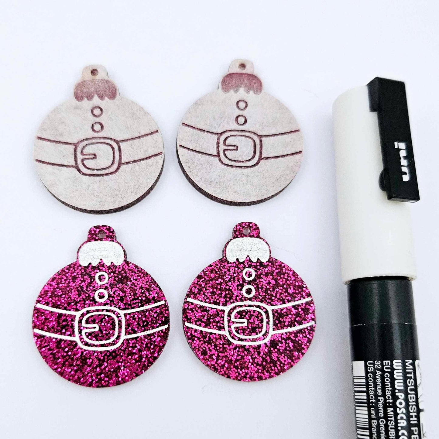 One pair of hot pink glitter Santa bauble dangle earring blanks paint filled white, alongside one unpainted pair with protective paper attached, pictured with a Posca paint pen