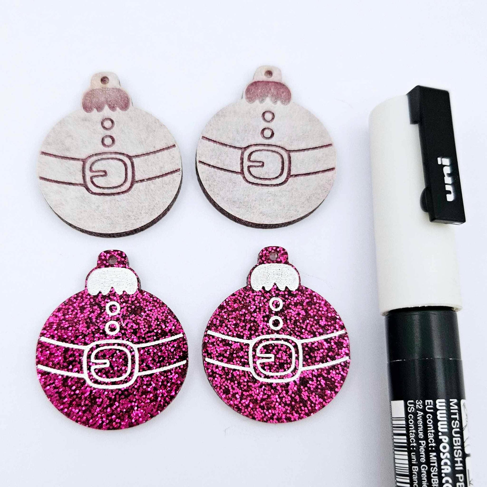 One pair of hot pink glitter Santa bauble dangle earring blanks paint filled white, alongside one unpainted pair with protective paper attached, pictured with a Posca paint pen