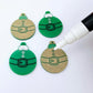 One pair of paint-filled solid green Santa bauble dangle blanks beside one unpainted pair with protective paper attached, with an uncapped Posca paint pen positioned ready to paint fill the engraved Santa belt details