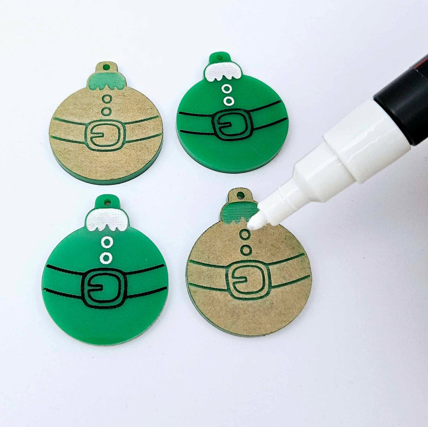 One pair of paint-filled solid green Santa bauble dangle blanks beside one unpainted pair with protective paper attached, with an uncapped Posca paint pen positioned ready to paint fill the engraved Santa belt details