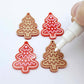 One pair of paint-filled solid red cookie Christmas tree dangle earring blanks and one unpainted pair with protective paper attached, with an uncapped Posca paint pen positioned ready to paint fill the engraved details
