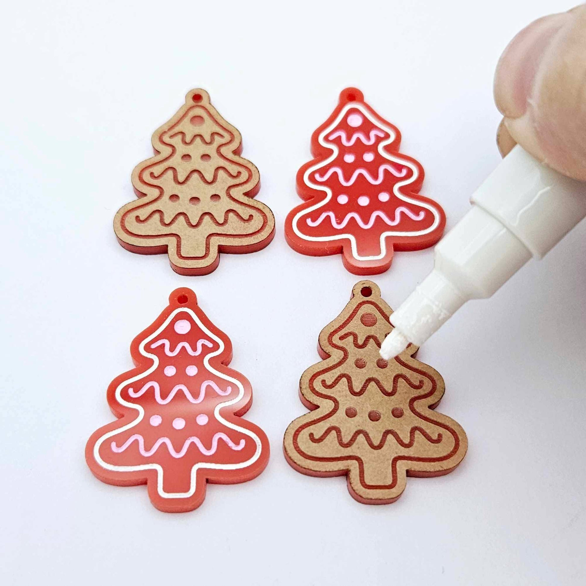 One pair of paint-filled solid red cookie Christmas tree dangle earring blanks and one unpainted pair with protective paper attached, with an uncapped Posca paint pen positioned ready to paint fill the engraved details