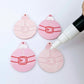 One pair of paint-filled pastel pink Santa bauble dangle blanks beside one unpainted pair with protective paper attached, with an uncapped Posca paint pen positioned ready to paint fill the engraved Santa belt details