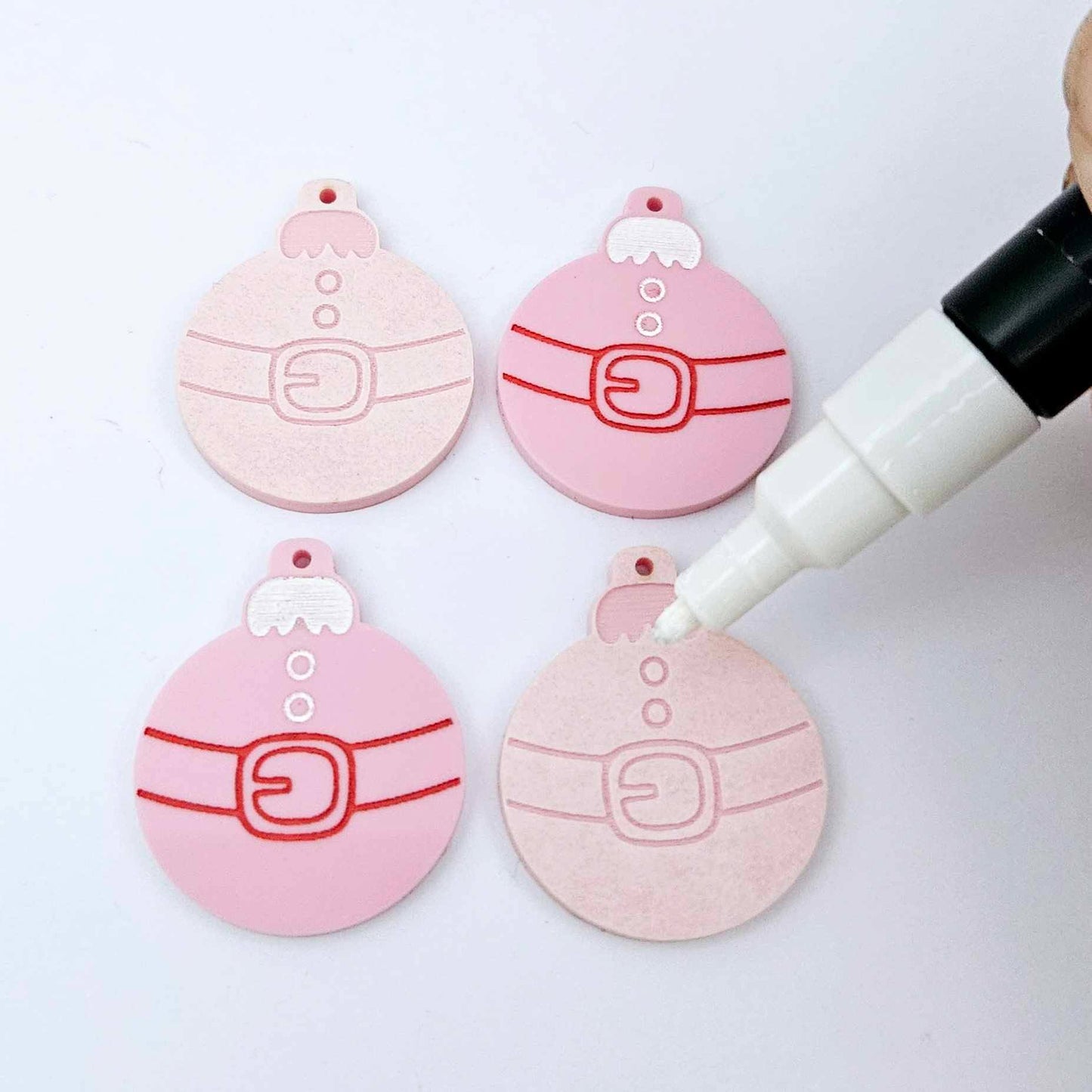 One pair of paint-filled pastel pink Santa bauble dangle blanks beside one unpainted pair with protective paper attached, with an uncapped Posca paint pen positioned ready to paint fill the engraved Santa belt details