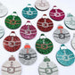Red, green, teal, silver and hot pink glitter Santa bauble dangle blanks paint filled, alongside unpainted pieces with protective paper attached