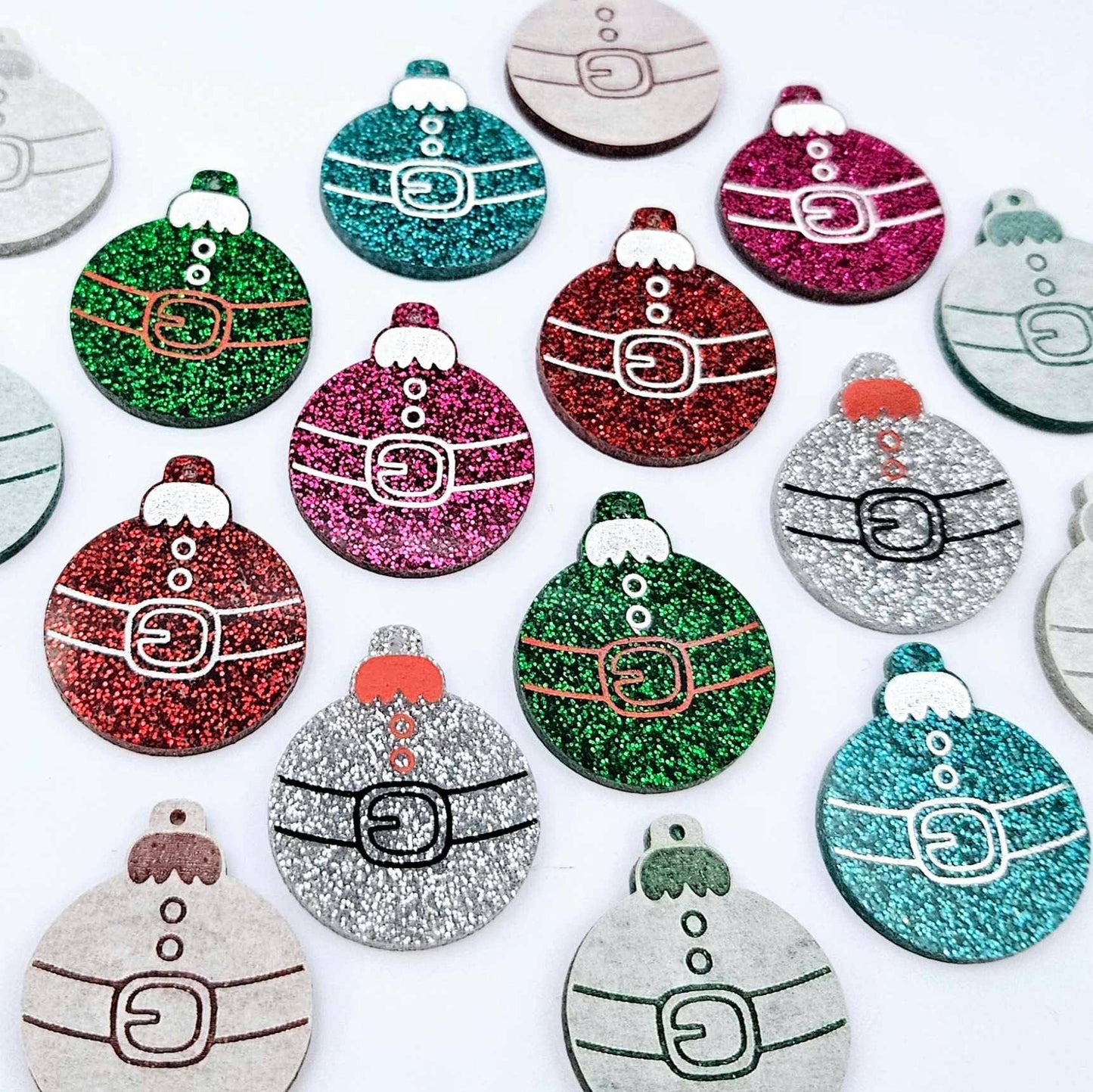 Red, green, teal, silver and hot pink glitter Santa bauble dangle blanks paint filled, alongside unpainted pieces with protective paper attached