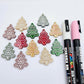 One pair of green, red and gold pearl marble cookie Christmas tree dangle earring blanks paint filled in light pink and white, alongside one unpainted pairs with protective paper attached, pictured with Posca paint pens