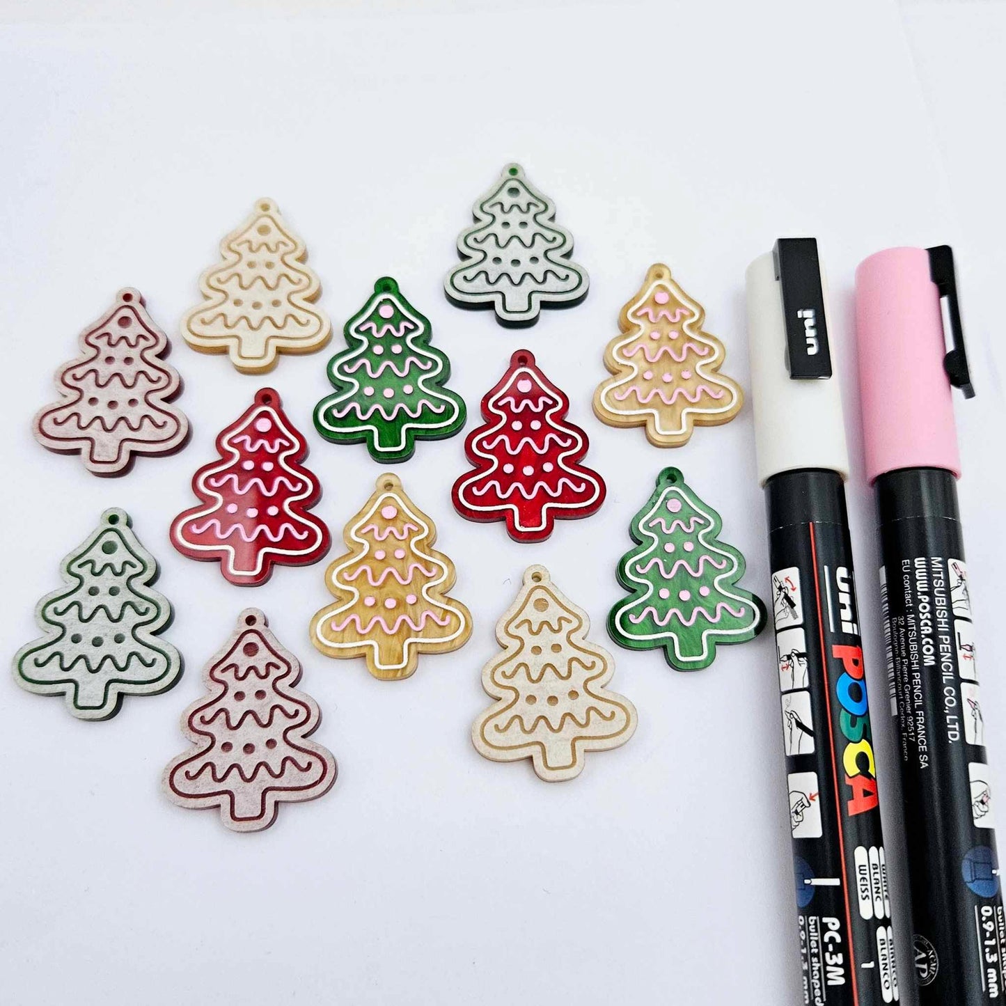One pair of green, red and gold pearl marble cookie Christmas tree dangle earring blanks paint filled in light pink and white, alongside one unpainted pairs with protective paper attached, pictured with Posca paint pens