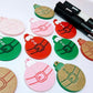 One paint-filled pair each of solid red, solid green and pastel pink Santa bauble dangle blanks, with additional unpainted blanks still covered with protective paper scattered around, showing the paint-fill possibilities.
