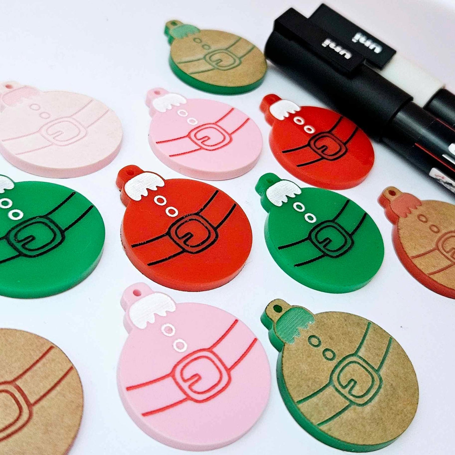 One paint-filled pair each of solid red, solid green and pastel pink Santa bauble dangle blanks, with additional unpainted blanks still covered with protective paper scattered around, showing the paint-fill possibilities.
