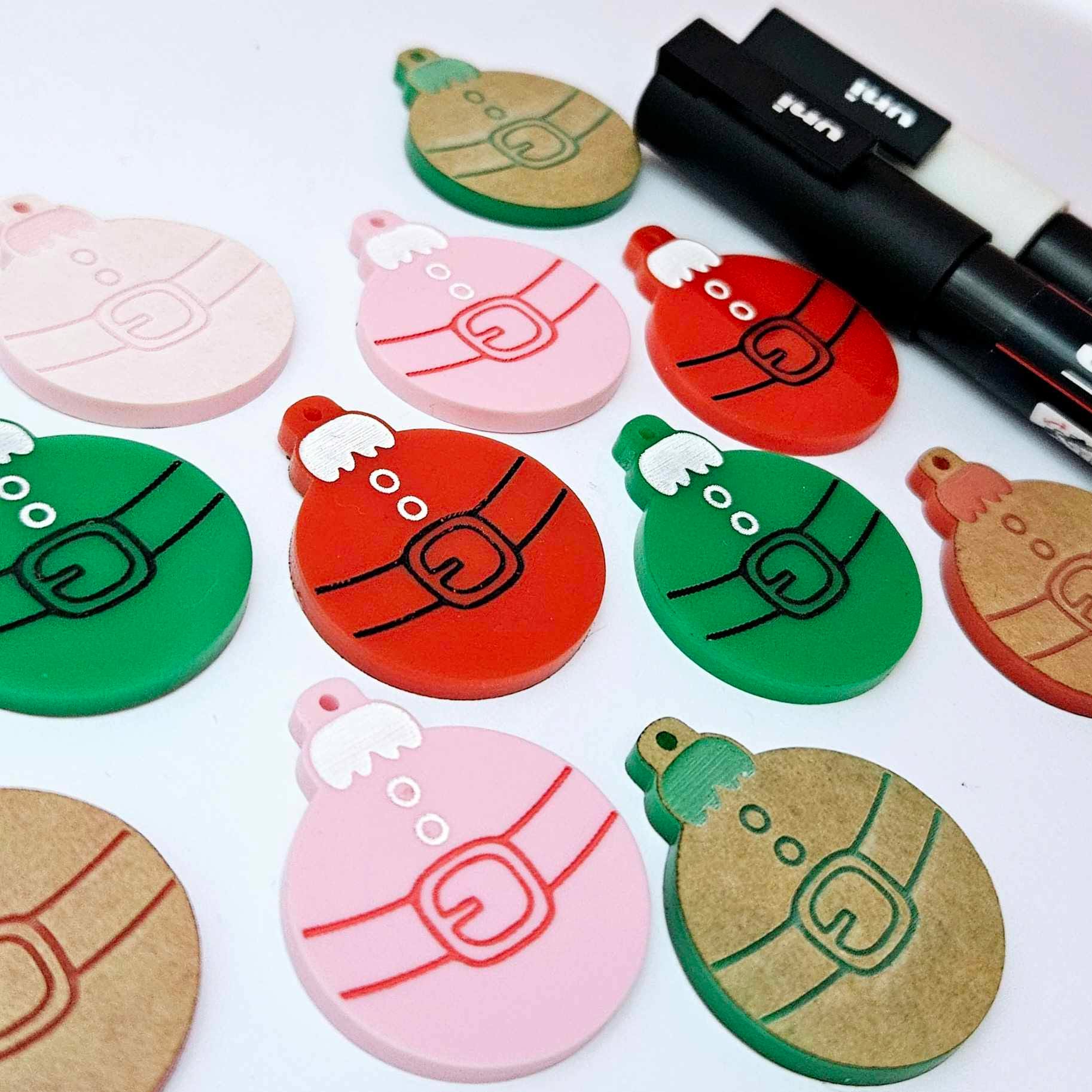 One paint-filled pair each of solid red, solid green and pastel pink Santa bauble dangle blanks, with additional unpainted blanks still covered with protective paper scattered around, showing the paint-fill possibilities.