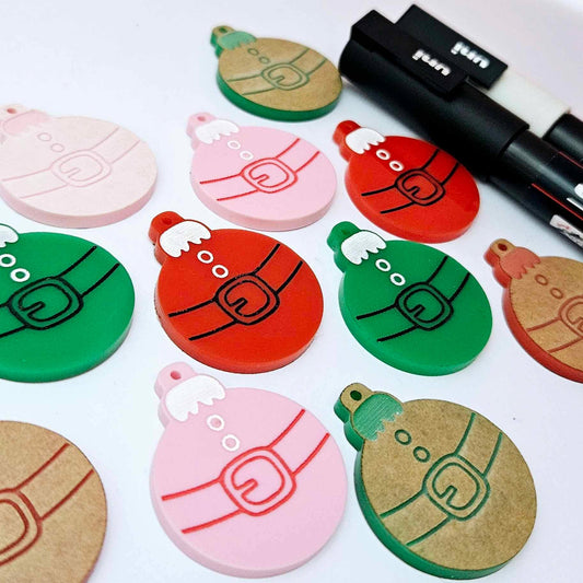 One paint-filled pair each of solid red, solid green and pastel pink Santa bauble dangle blanks, with additional unpainted blanks still covered with protective paper scattered around, showing the paint-fill possibilities.