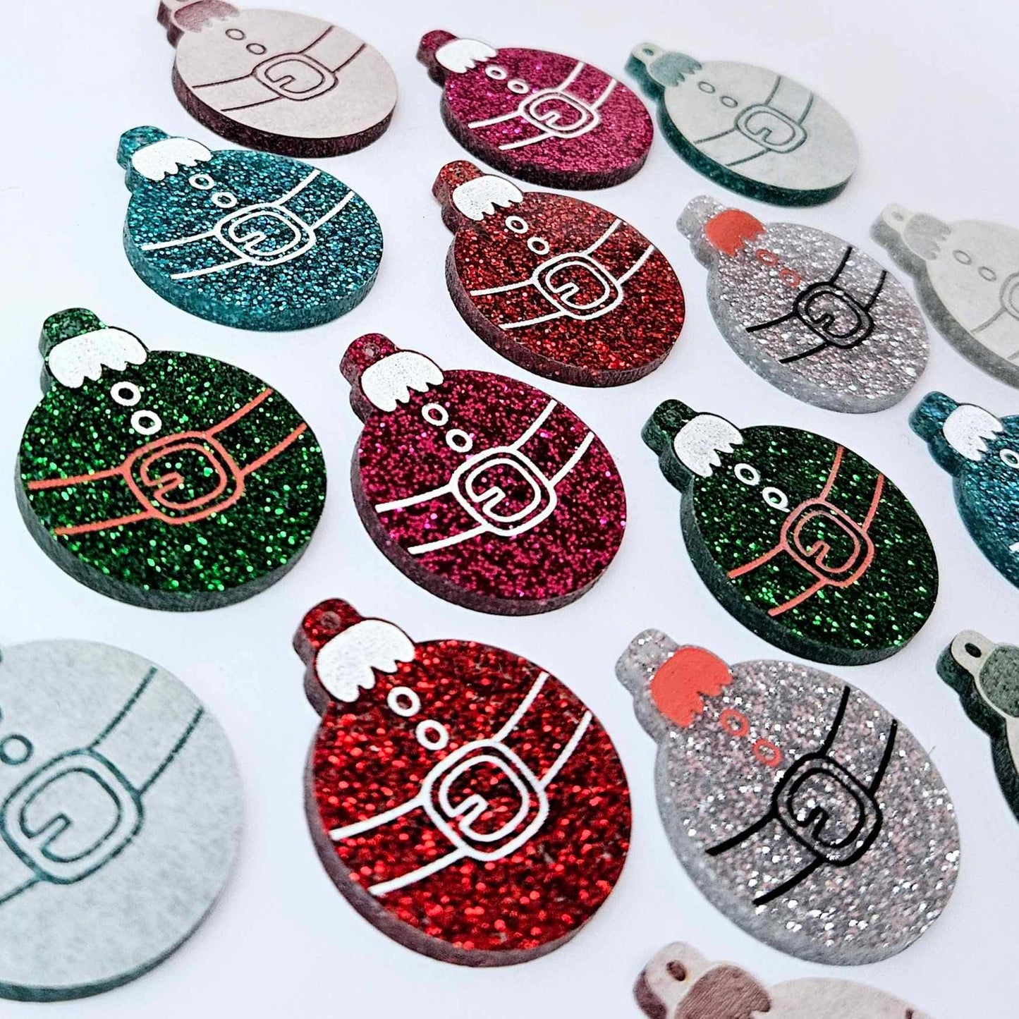 Red, green, teal, silver and hot pink glitter Santa bauble dangle blanks paint filled, alongside unpainted pieces with protective paper attached