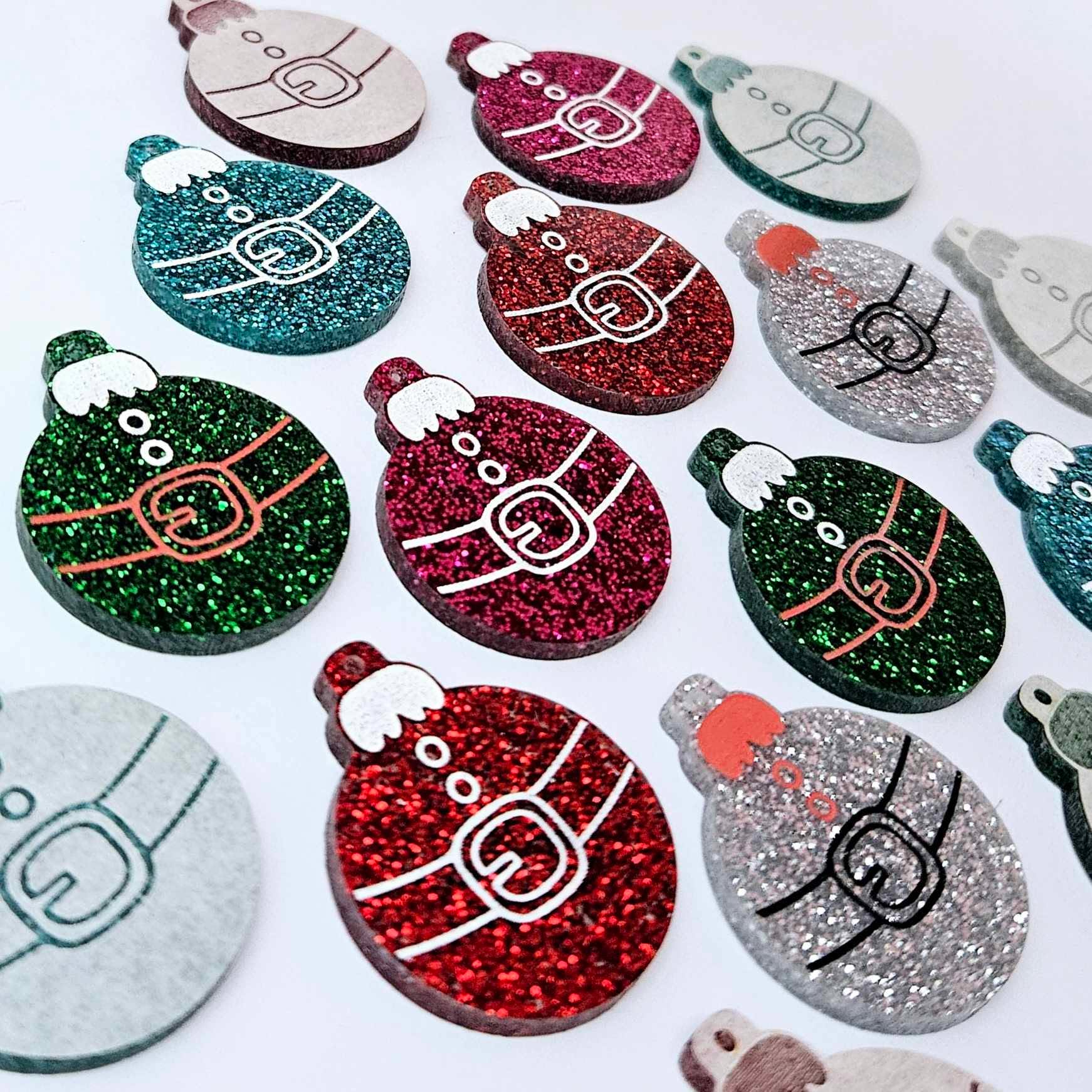 Red, green, teal, silver and hot pink glitter Santa bauble dangle blanks paint filled, alongside unpainted pieces with protective paper attached