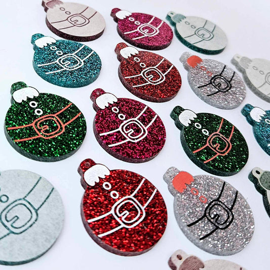 Red, green, teal, silver and hot pink glitter Santa bauble dangle blanks paint filled, alongside unpainted pieces with protective paper attached