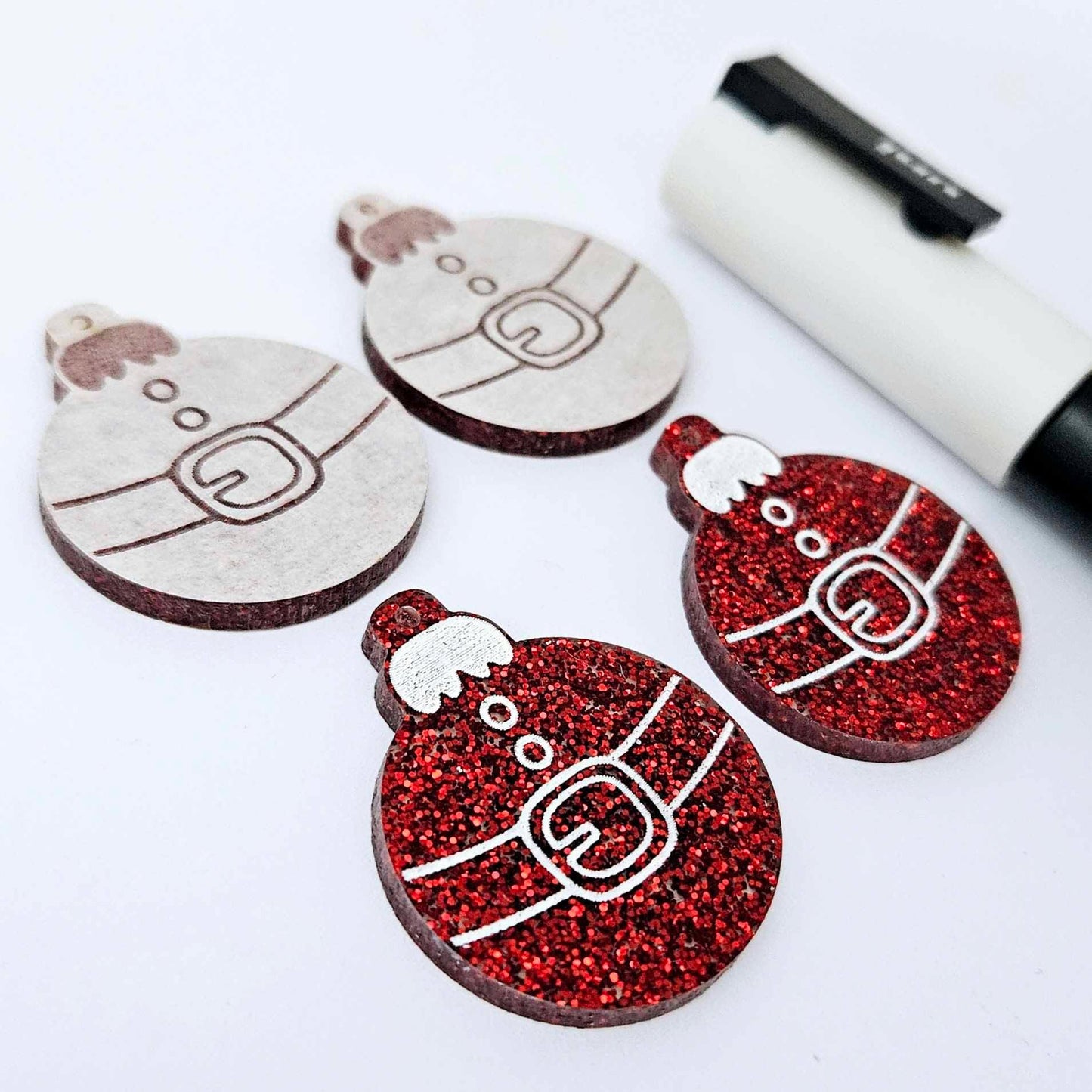One pair of red glitter Santa bauble dangle blanks paint filled white, alongside one unpainted pair with protective paper attached, pictured with Posca paint pens