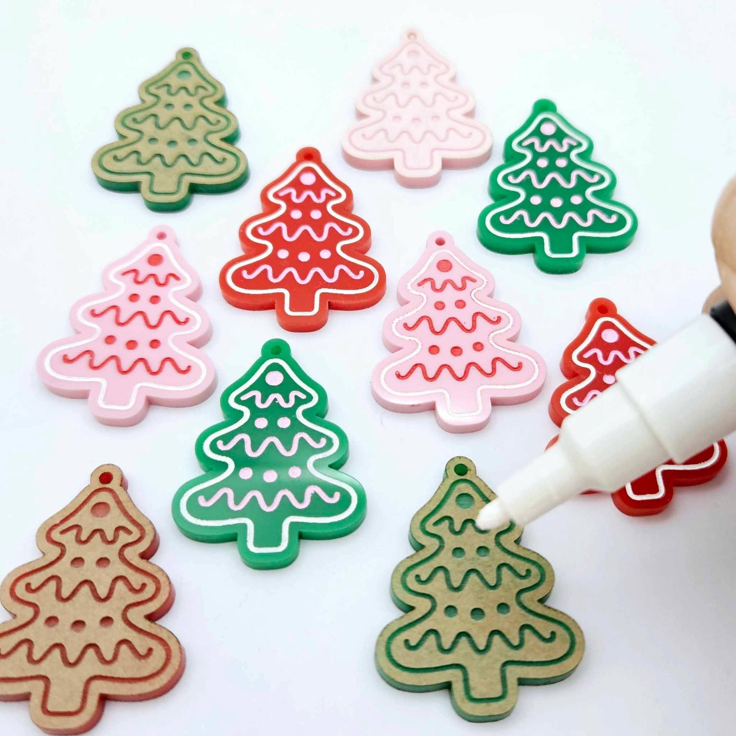 Cookie Christmas tree dangle earring blanks in solid red, solid green and pastel pink, showing a mix of paint-filled and unpainted pieces with a Posca paint pen positioned ready to paint fill the engraved details