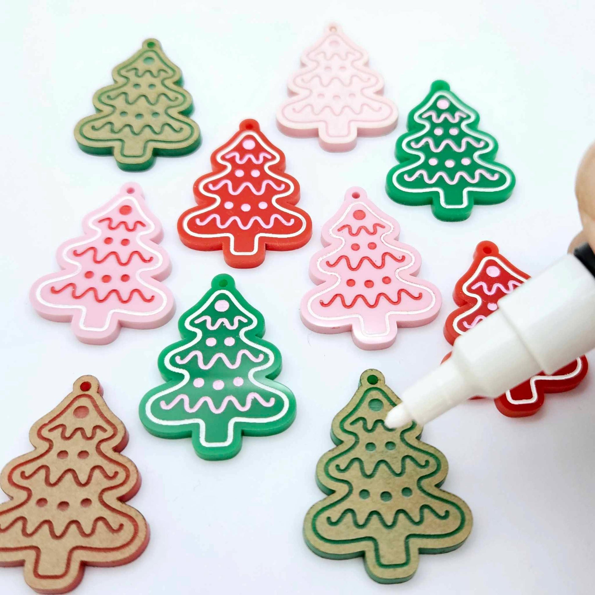 Cookie Christmas tree dangle earring blanks in solid red, solid green and pastel pink, showing a mix of paint-filled and unpainted pieces with a Posca paint pen positioned ready to paint fill the engraved details