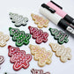 One pair of green, red and gold pearl marble cookie Christmas tree dangle earring blanks paint filled in light pink and white, alongside one unpainted pairs with protective paper attached, pictured with Posca paint pens