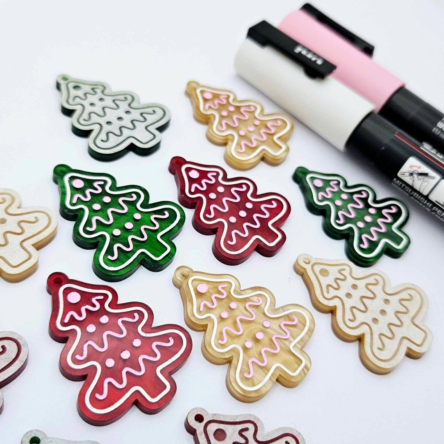 One pair of green, red and gold pearl marble cookie Christmas tree dangle earring blanks paint filled in light pink and white, alongside one unpainted pairs with protective paper attached, pictured with Posca paint pens