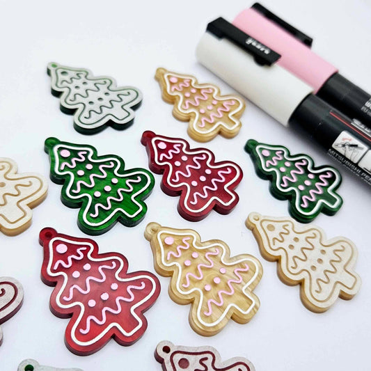 One pair of green, red and gold pearl marble cookie Christmas tree dangle earring blanks paint filled in light pink and white, alongside one unpainted pairs with protective paper attached, pictured with Posca paint pens