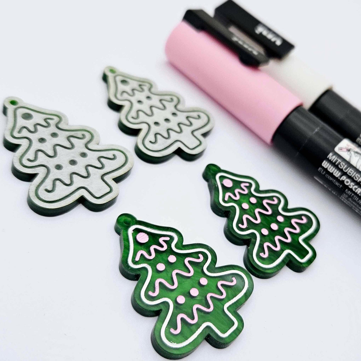 One pair of green marble cookie Christmas tree dangle blanks paint filled in light pink and white, alongside one unpainted pair with protective paper attached, pictured with Posca paint pens